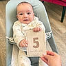 baby, infant, milestone_card, 5_months_old, baby_seat, smiling, cozy_clothing, hand, wooden_floor, indoor, child, portrait, cute, happy, person, seat_belt, home, flooring, young_child, holding