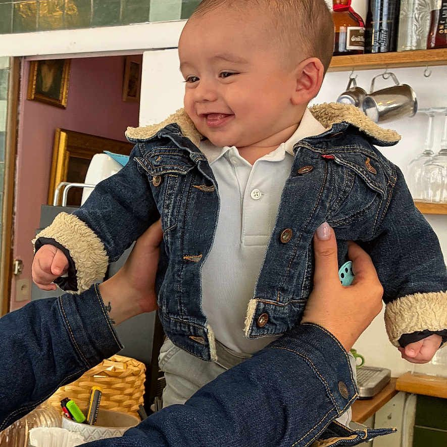 Ange a rejoint le concours — aidez-le/la à gagner de superbes lots ! baby, denim_jacket, smiling, child, indoor, kitchen, wooden_table, adult_hands, bottle, jar, shelf, cupboard, white_socks, casual_clothing, happy, portrait, person, holding, table, cozy