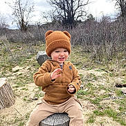 Caden is registered to the contest to win money with this photo: toddler, child, hat, knit_hat, bear_ears, brown_clothing, hoodie, pants, black_shoes, outdoor, tree_stump, grass, dry_brush, trees, cloudy_sky, nature, playful, smiling, sitting, cute
