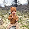 toddler, child, hat, knit_hat, bear_ears, brown_clothing, hoodie, pants, black_shoes, outdoor, tree_stump, grass, dry_brush, trees, cloudy_sky, nature, playful, smiling, sitting, cute