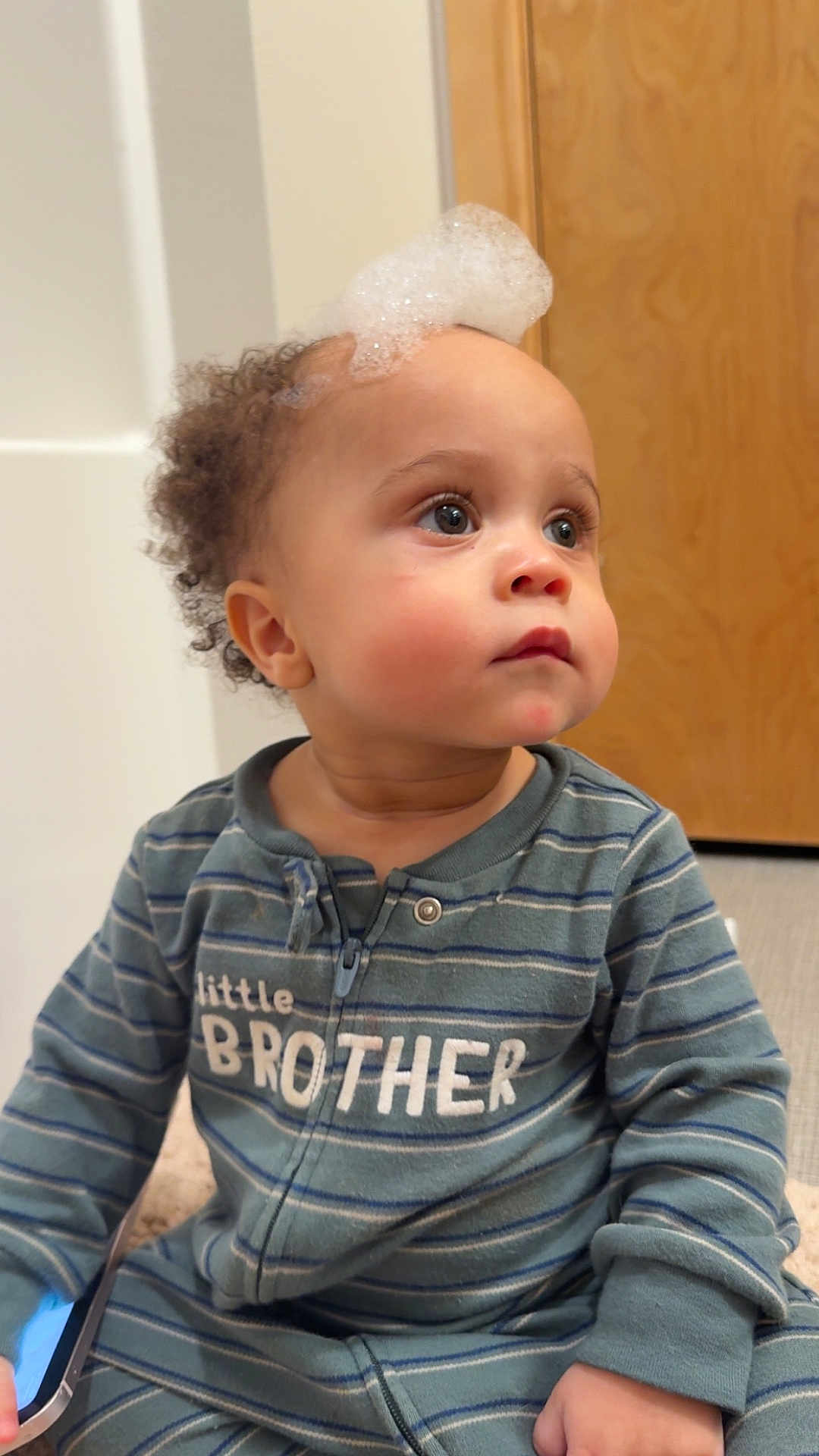 Siylas is registered to the contest to win money with this photo: toddler, child, baby, striped_pajamas, face, curly_hair, bubbles, foam, indoor, floor, door, long_sleeve, zipper, blue, green, expression, sitting, hand, looking_away, clothing