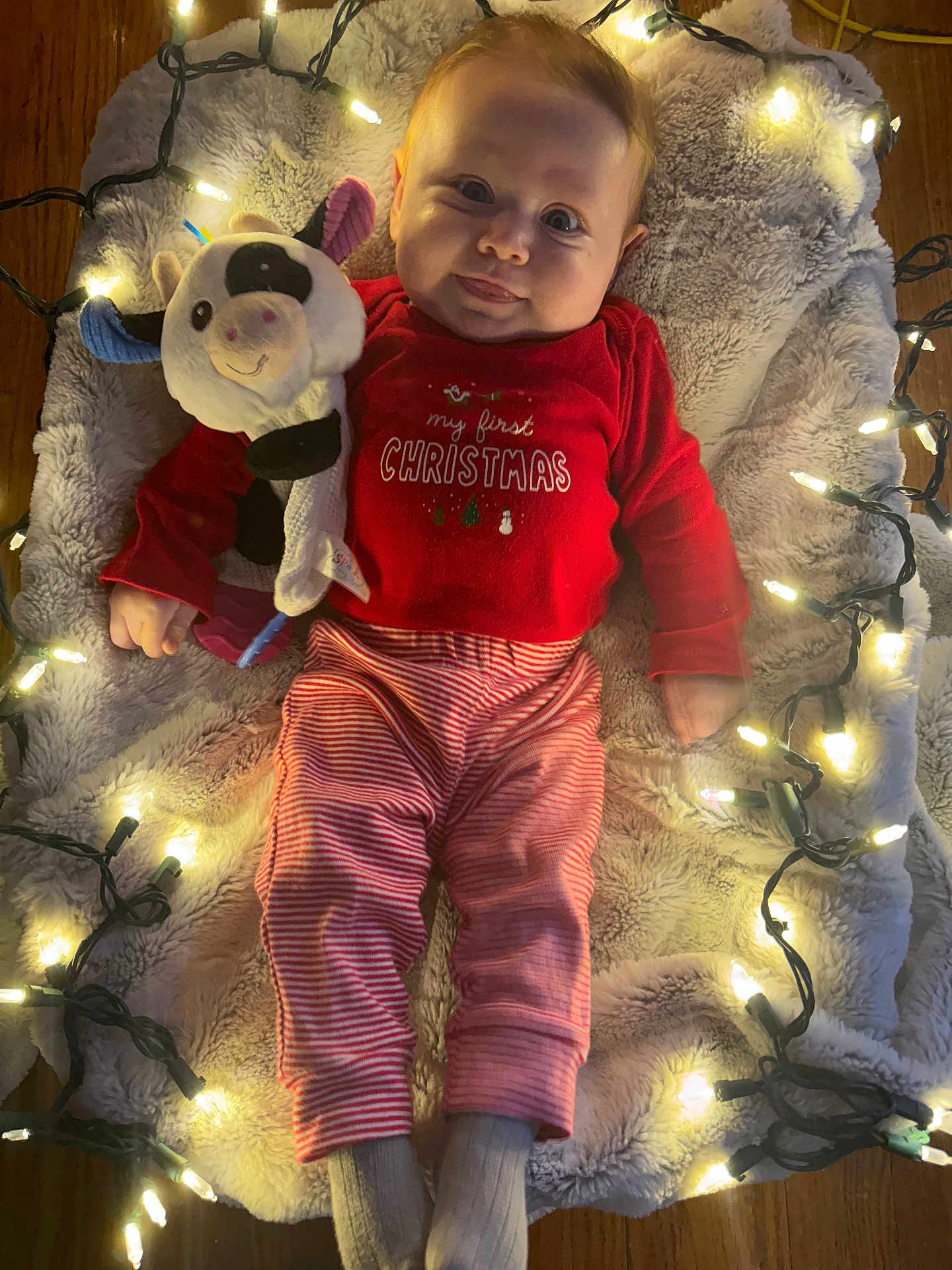 Carina is registered to the contest to win money with this photo: baby, infant, smile, christmas, lights, blanket, plush_toy, stuffed_animal, pajamas, red_clothing, striped_pants, socks, hardwood_floor, cozy, holiday, portrait, lying_down, fur_rug, fairy_lights, cute
