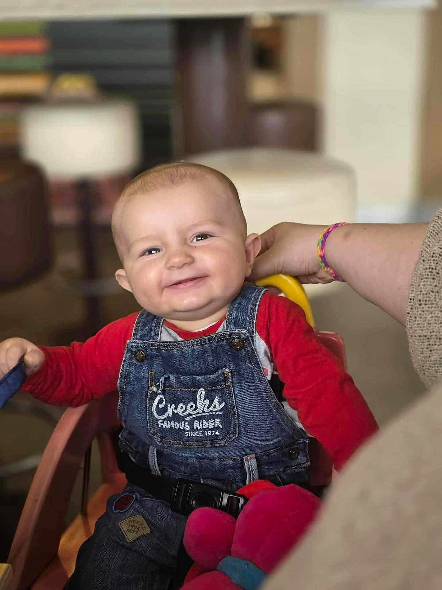Helio participe au concours pour gagner de l'argent avec cette photo : baby, child, smiling, denim_overalls, red_shirt, high_chair, indoor, hand, person, cute, happy, seated, furniture, toy, plush_toy, portrait, face, young_child, casual_clothing, support