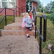 Kenyatta is registered to the contest to win money with this photo: child, girl, backpack, steps, handrail, building, brick, outdoor, sidewalk, grass, trees, shoes, socks, clothing, person, daylight, smile, hair_beads, casual, ready_for_school