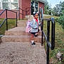 child, girl, backpack, steps, handrail, building, brick, outdoor, sidewalk, grass, trees, shoes, socks, clothing, person, daylight, smile, hair_beads, casual, ready_for_school