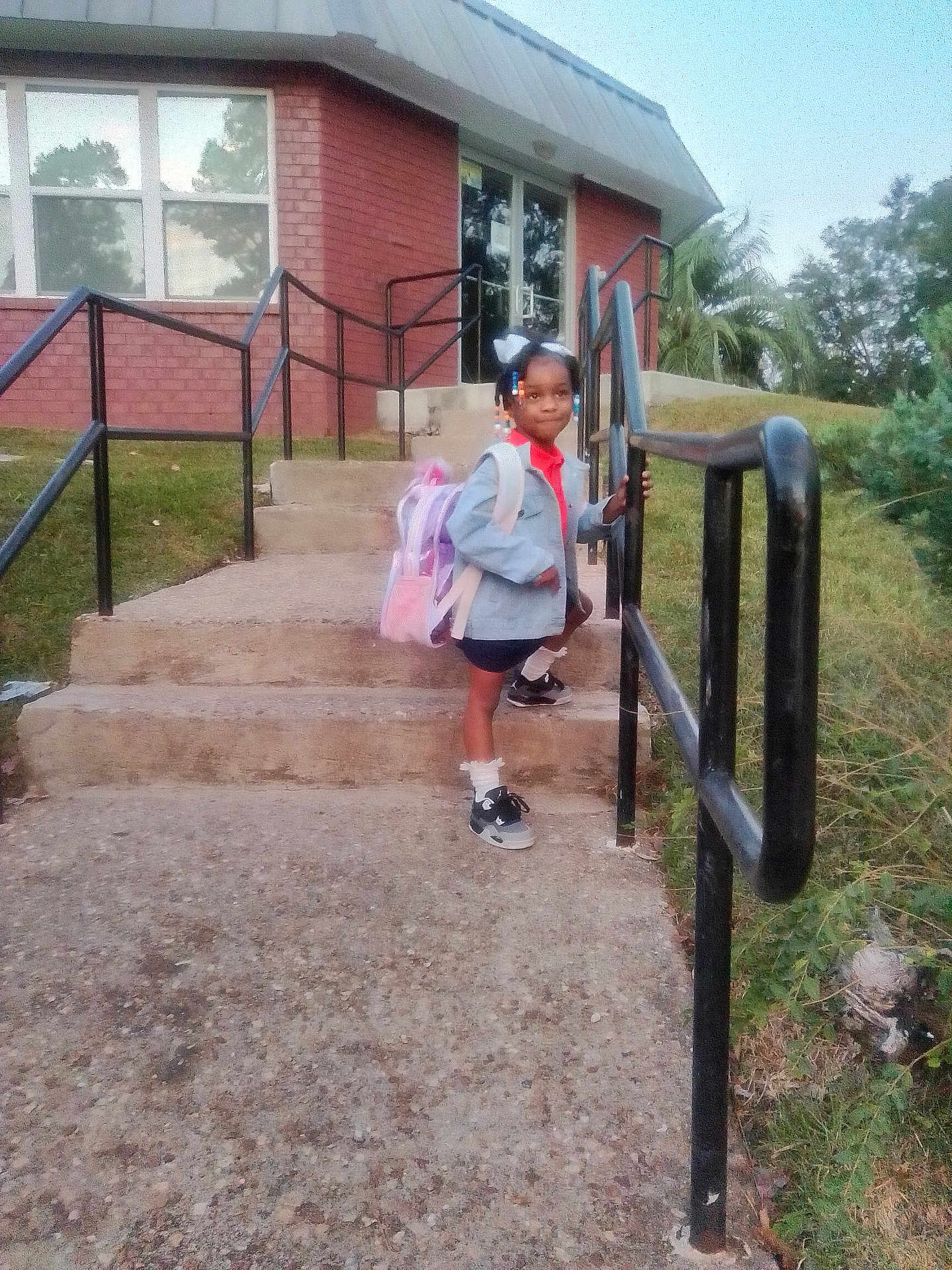Kenyatta is registered to the contest to win money with this photo: child, girl, backpack, steps, handrail, building, brick, outdoor, sidewalk, grass, trees, shoes, socks, clothing, person, daylight, smile, hair_beads, casual, ready_for_school