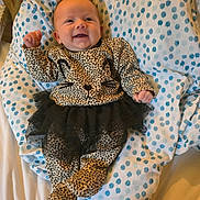 Samantha is registered to the contest to win money with this photo: baby, infant, smiling, leopard_print, onesie, tutu, black_tutu, blanket, polka_dot, blue, white, happy, child, lying_down, cute, fist_raised, face, person, clothing, bed