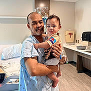 Hamlin is registered to the contest to win money with this photo: baby, child, father, parent, hospital, hospital_room, medical_equipment, patient, infant, holding, family, portrait, indoors, bed, hospital_gown, medical_tubing, bandage, caregiver, sitting, photograph