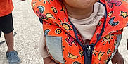 Ezekiel a rejoint le concours — aidez-le/la à gagner de superbes lots ! toddler, child, life_jacket, orange, smile, outdoor, gravel, playful, sun_pattern, stars, clothing, person, footwear, red_shoes, happy, curly_hair, zipper, straps, safety, standing