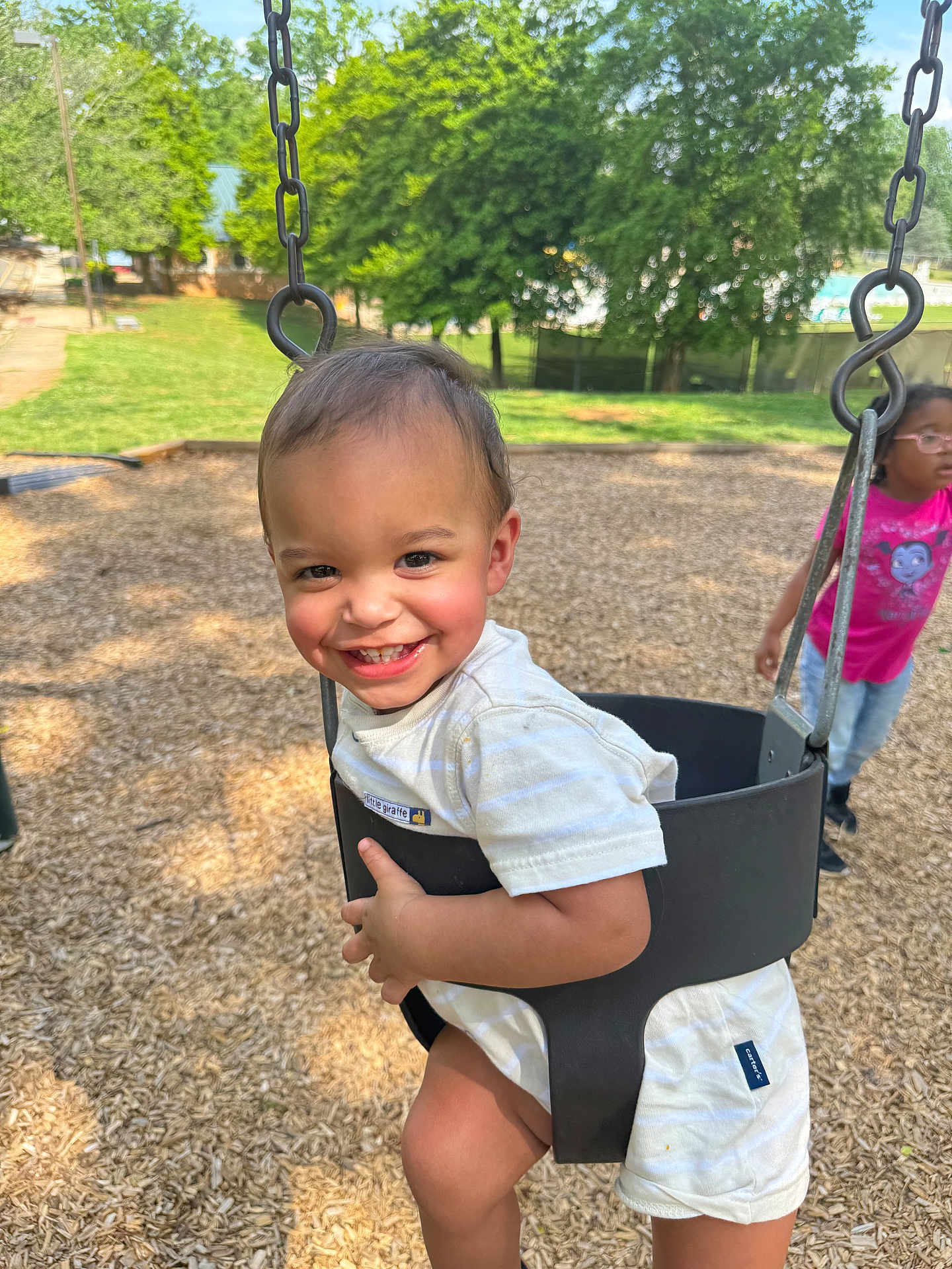 Roman joined the competition — help win amazing prizes! toddler, swing, playground, child, smile, outdoor, daylight, trees, woodchips, happy, black_swing_seat, grass, person, glasses, pink_shirt, shorts, sunlight, park, play, fun
