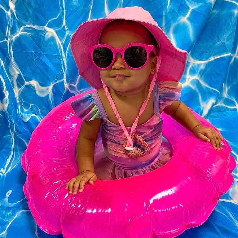 Miabelle is registered to the contest to win money with this photo: accessories, baby, blue_background, child, colorful, cute, fashion, fun, indoor, inflatable_ring, pink_hat, playful, portrait, smiling, summer, sunglasses, swimwear, toddler, vacation, water_pattern
