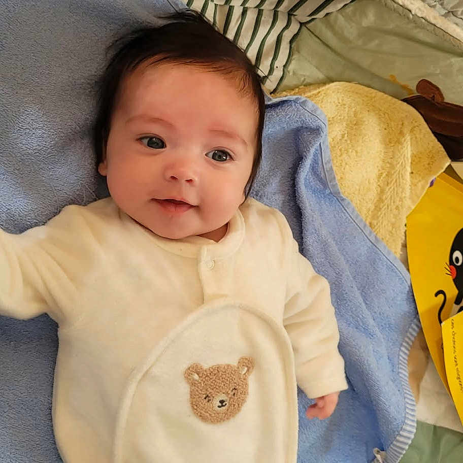 Léa participe au concours pour gagner de l'argent avec cette photo : baby, infant, smile, onesie, bear_patch, blanket, towel, blue_blanket, yellow_book, book_cover, dark_hair, cheek, eyes, lying_down, portrait, cozy, soft_toys, hand, nursery, cute