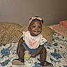 baby, child, infant, smile, headband, bib, denim_shorts, toy, sandals, bed, pillow, blanket, floral_bedspread, indoor, person, cute, sitting, happy, young_child, cozy