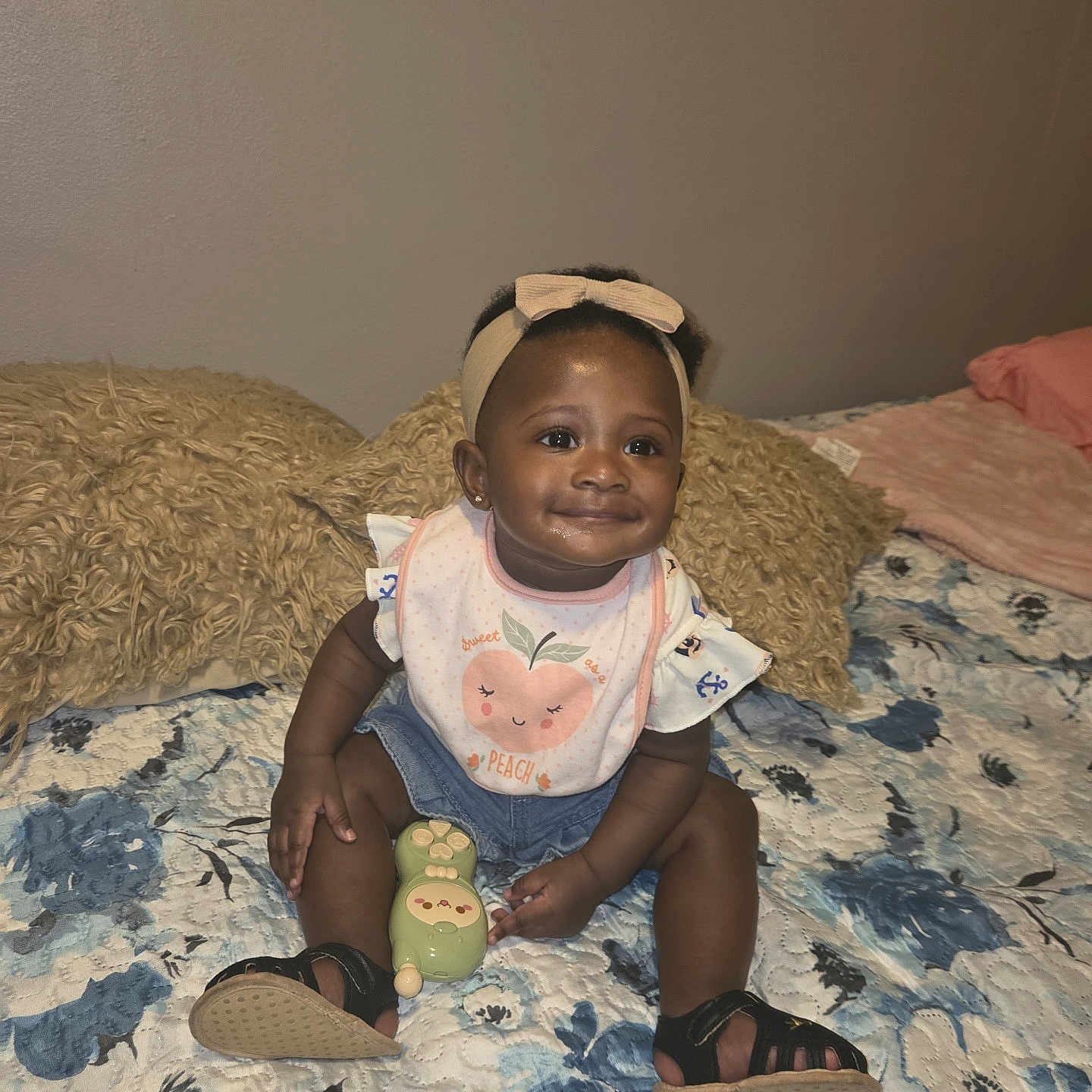Zunaisha is registered to the contest to win money with this photo: baby, child, infant, smile, headband, bib, denim_shorts, toy, sandals, bed, pillow, blanket, floral_bedspread, indoor, person, cute, sitting, happy, young_child, cozy