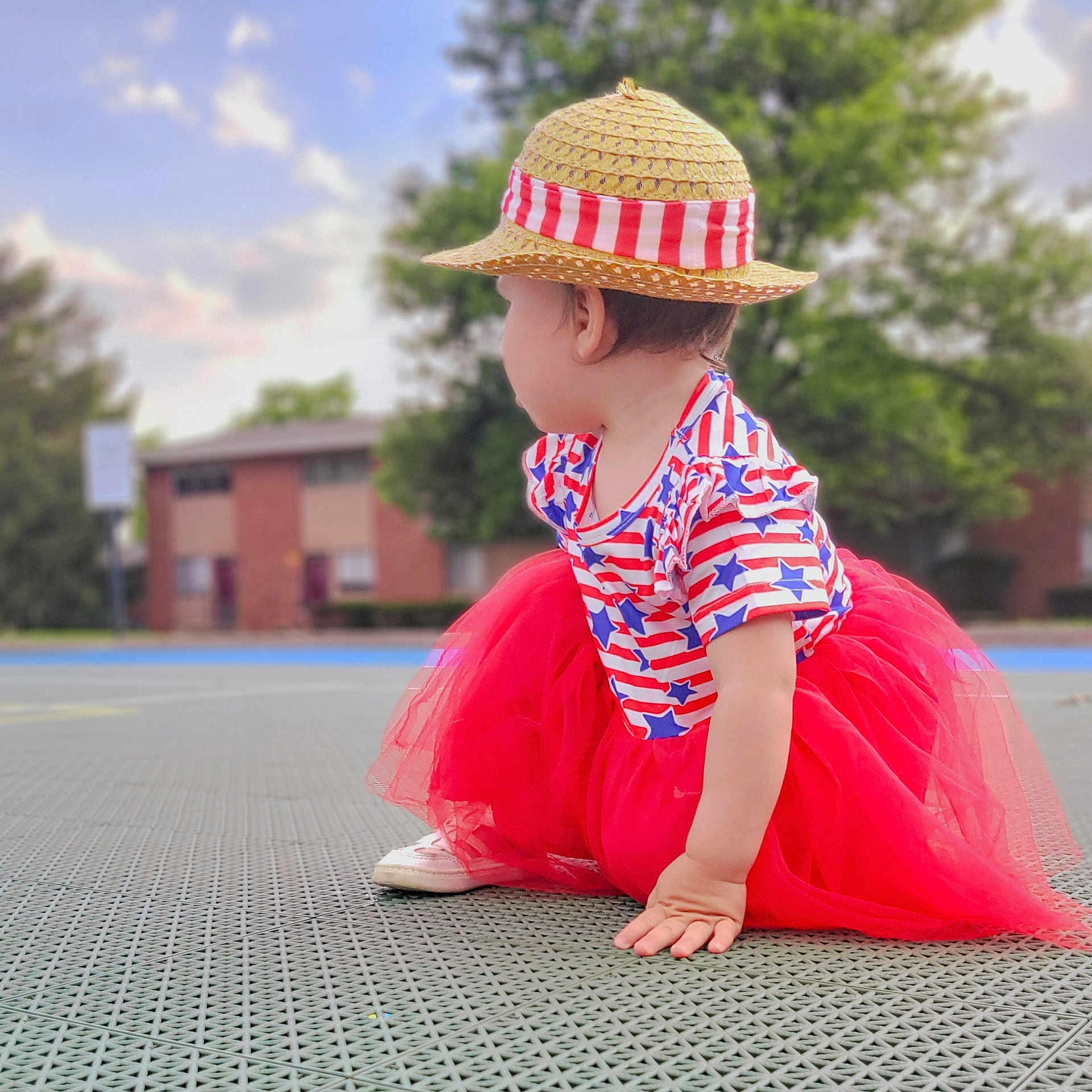 Freyja is registered to the contest to win money with this photo: toddler, child, hat, straw_hat, red_tutu, red_skirt, patriotic_clothing, stars_and_stripes, outdoor, daylight, crouching, side_view, grass, trees, building, footwear, cute, fashion, summer, playground