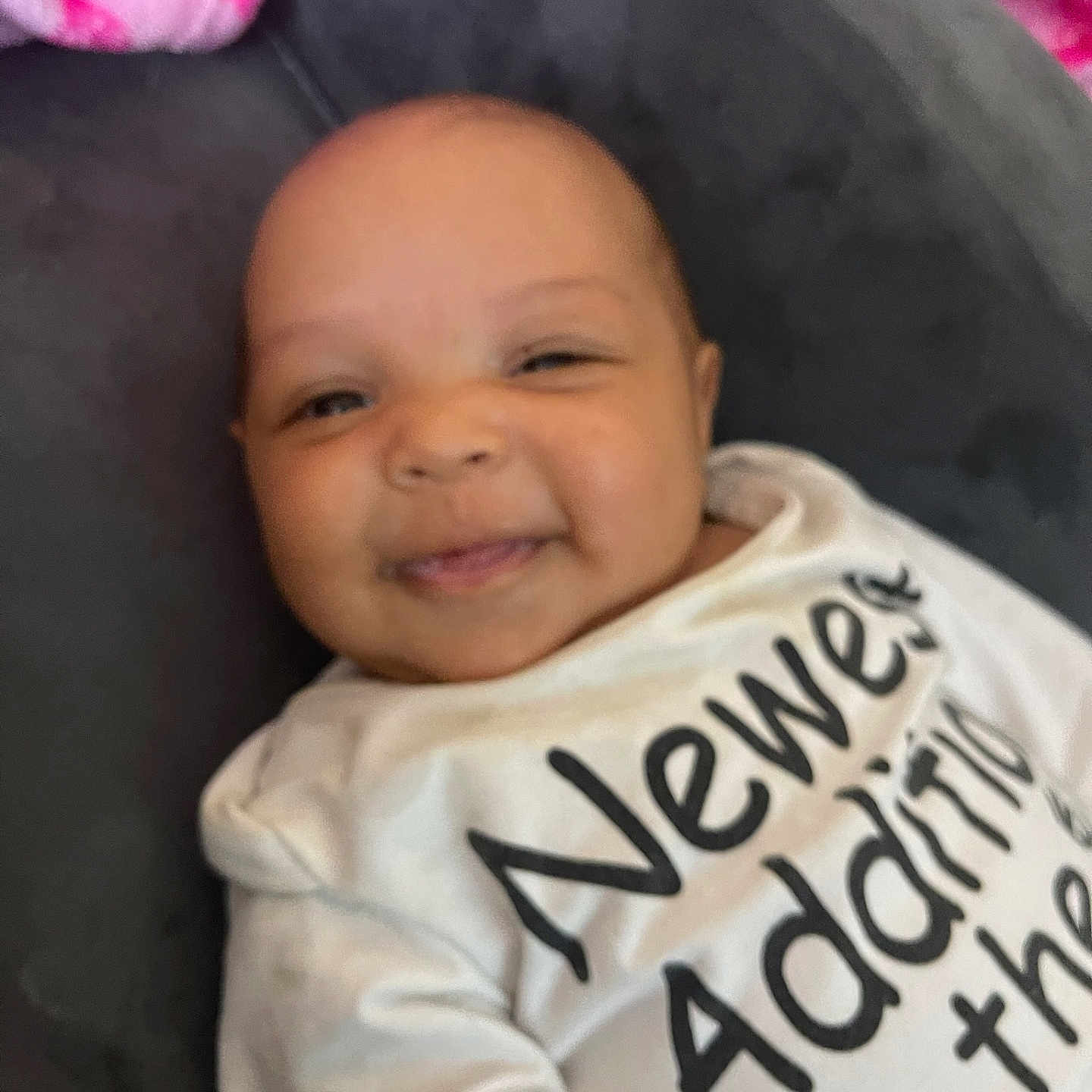 Inez joined the competition — help win amazing prizes! baby, child, clothing, cozy, cushion, cute, face, gray_cushion, happy, indoors, infant, lying_down, person, pink_blanket, portrait, smiling, soft_texture, text_on_clothing, white_shirt, young