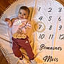 baby, child, smiling, pink_clothing, adidas, white_shoes, wooden_sign, wooden_wreath, round_blanket, numbers, text, floor, celebration, milestone, indoors, person, happy, portrait, sitting, cute
