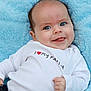 baby, blue_blanket, blue_eyes, child, closeup, cozy, cute, face, happy, infant, lying_down, person, portrait, red_heart, smiling, soft_blanket, text_on_clothing, tongue_out, white_shirt, young