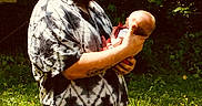 Preston is registered to the contest to win money with this photo: man, baby, outdoor, grass, nature, person, holding, cap, beard, shirt, tie_dye, jeans, sunlight, greenery, care, affection, portrait, father, child, tenderness