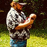 man, baby, outdoor, grass, nature, person, holding, cap, beard, shirt, tie_dye, jeans, sunlight, greenery, care, affection, portrait, father, child, tenderness