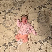 Alaiyah is registered to the contest to win money with this photo: baby, infant, smiling, pink_clothing, bedspread, keys_pattern, script_text, lying_down, raised_arm, white_socks, blonde_hair, cute, indoor, portrait, child, happy, soft_lighting, fabric, cozy, toddler