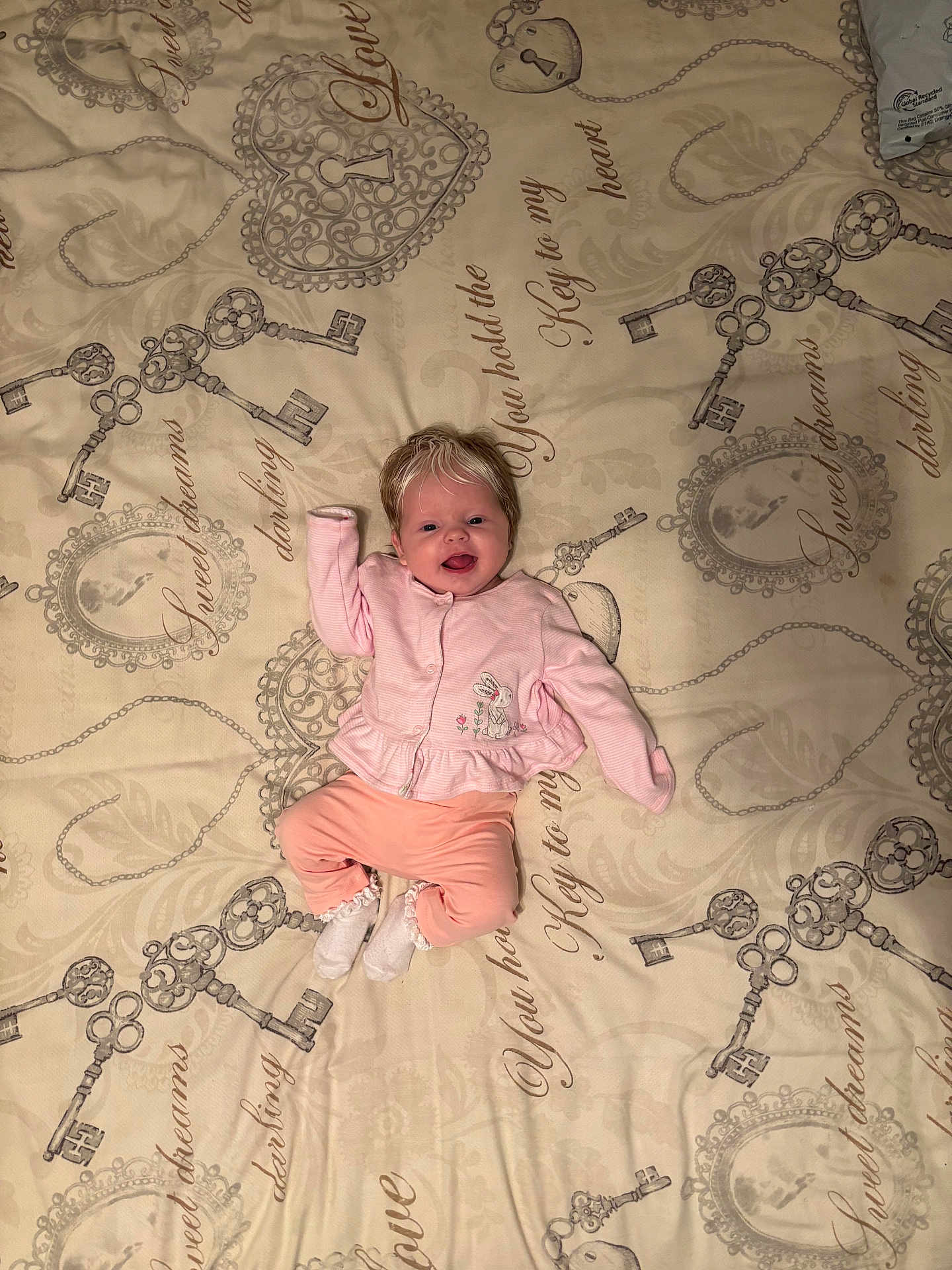 Alaiyah is registered to the contest to win money with this photo: baby, infant, smiling, pink_clothing, bedspread, keys_pattern, script_text, lying_down, raised_arm, white_socks, blonde_hair, cute, indoor, portrait, child, happy, soft_lighting, fabric, cozy, toddler