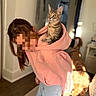 cat, tabby_cat, hoodie, pink_hoodie, person, indoor, dog, curious_cat, light, warm_light, wood_floor, carpet, furniture, pet, domestic_animal, animal, cozy, playful, home, casual_clothing