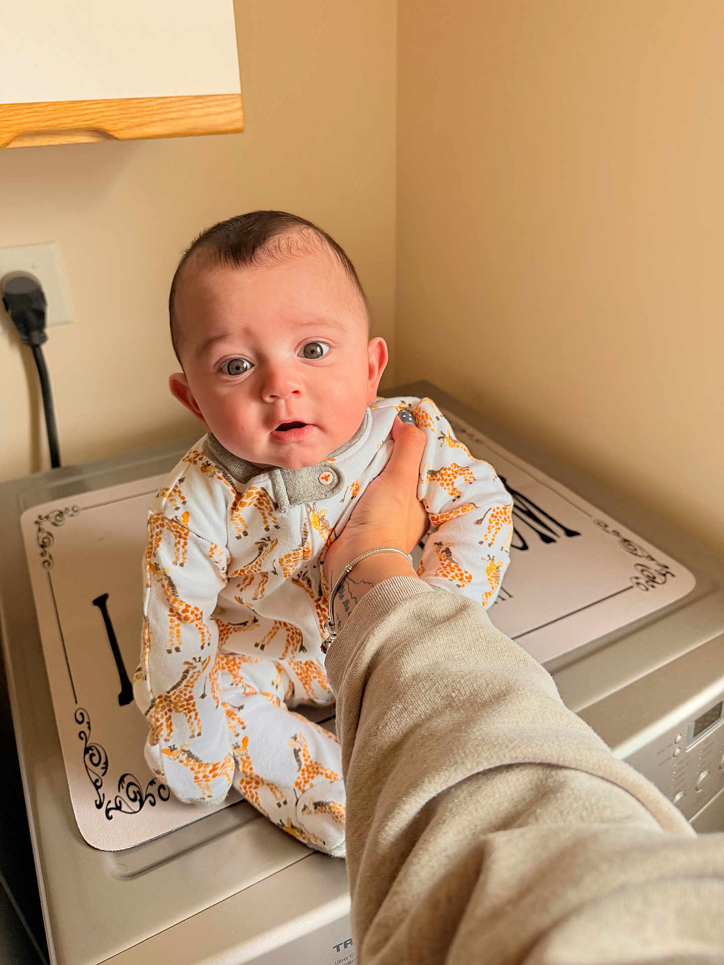 Michael is registered to the contest to win money with this photo: baby, infant, giraffe_pajamas, washing_machine, hand, person, indoor, curious, expression, clothing, appliance, face, child, cute, home, sleepsuit, holding, young, portrait, light