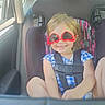 child, toddler, smiling, sunglasses, heart_shaped_sunglasses, car, car_seat, safety_harness, vehicle_interior, road_trip, blonde_hair, blue_dress, checkered_clothing, portrait, sitting, indoor, daylight, happy, family, travel