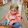 child, toddler, shopping_cart, hardware_store, store_aisle, orange_cart, curly_hair, blond_hair, pouting, portrait, sitting, indoors, retail, shelves, merchandise, shopping, hoodie, jeans, facial_expression, photography
