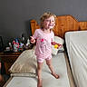 child, toddler, girl, smiling, happy, portrait, bedroom, bed, mattress, pajamas, pink_clothing, barefoot, toy, thumbs_up, wooden_headboard, pillows, bedside_table, indoors, home, daylight