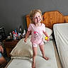 child, girl, toddler, smiling, happy, portrait, bedroom, mattress, bed, barefoot, pink_clothing, pajamas, unicorn_shirt, toy, yellow_object, wooden_headboard, pillow, nightstand, indoor, family_photo