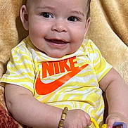 Mylan joined the competition — help win amazing prizes! baby, smiling, toy, yellow_shirt, nike_logo, bracelet, blanket, child, happy, cute, indoor, face, person, infant, playful, holding, colorful, small_hands, portrait, seated