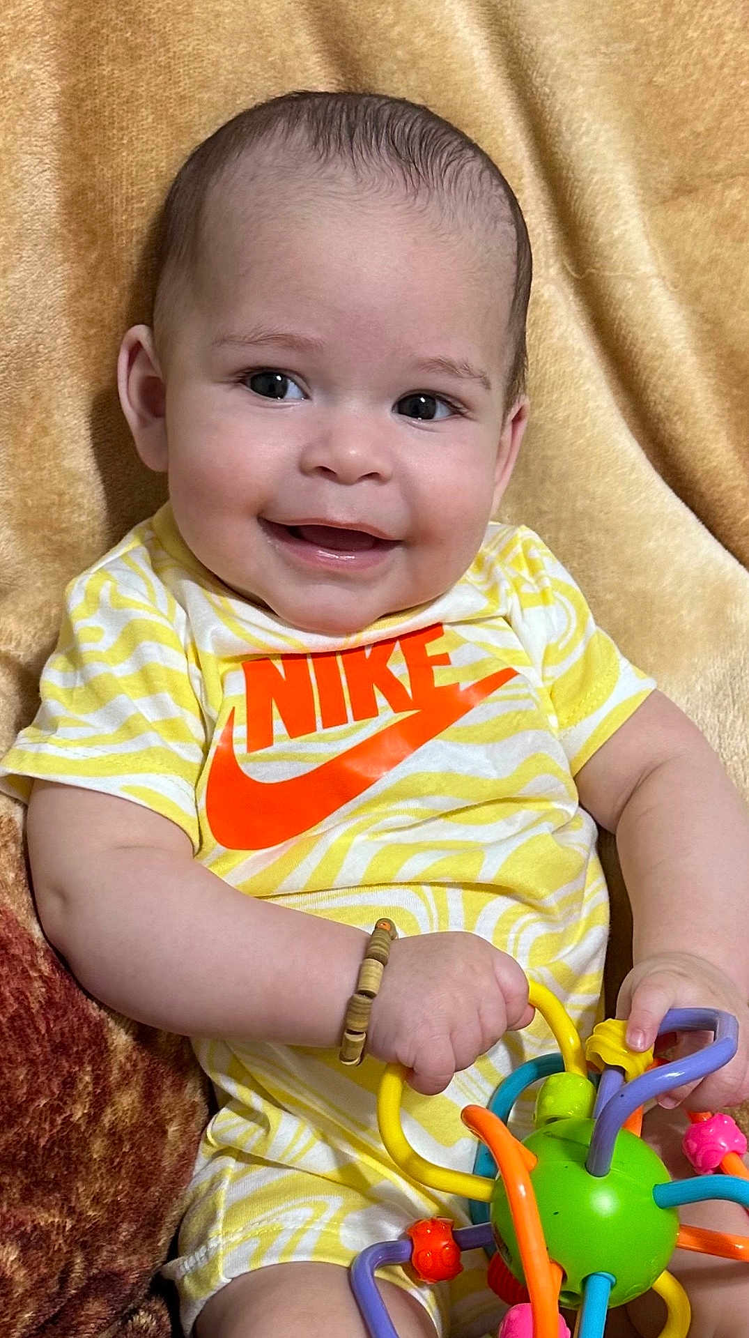 Mylan joined the competition — help win amazing prizes! baby, smiling, toy, yellow_shirt, nike_logo, bracelet, blanket, child, happy, cute, indoor, face, person, infant, playful, holding, colorful, small_hands, portrait, seated