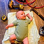 James is registered to the contest to win money with this photo: baby, infant, child, floor, wooden_floor, tools, drill, hammer, tape_measure, hose, onesie, boots, carpet, smiling, portrait, indoor, cute, playful, person, clothing