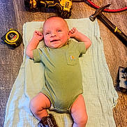 James joined the competition — help win amazing prizes! baby, infant, smile, onesie, boots, cowboy_boots, blanket, tools, drill, hammer, tape_measure, wood_floor, flooring, laying_down, happy, child, indoor, person, cute, small