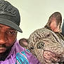 man, dog, french_bulldog, cap, hoodie, purple_clothing, pet, selfie, indoor, closeup, wrinkles, face, portrait, expression, companion, animal, casual, beard, headwear, friendship