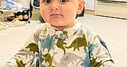 Davud joined the competition — help win amazing prizes! toddler, child, high_chair, dinosaur_pajamas, indoors, kitchen, shoes, stroller, curious_expression, baby, face, person, furniture, home, floor, cabinet, tray, hand, sitting, young_child