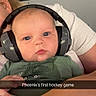 baby, infant, child, face, portrait, headphones, earmuffs, hockey_game, sports_arena, spectator, green_shirt, adult, parent, indoor, seating, close_up, wide_eyed, calm, text_overlay, selfie