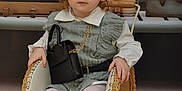 Aliya participe au concours pour gagner de l'argent avec cette photo : toddler, child, red_hair, curly_hair, wicker_stroller, black_boots, gray_dress, white_shirt, handbag, gold_chain, vintage, wooden_trunks, candles, indoor, portrait, cute, fashion, seated, furniture, decor