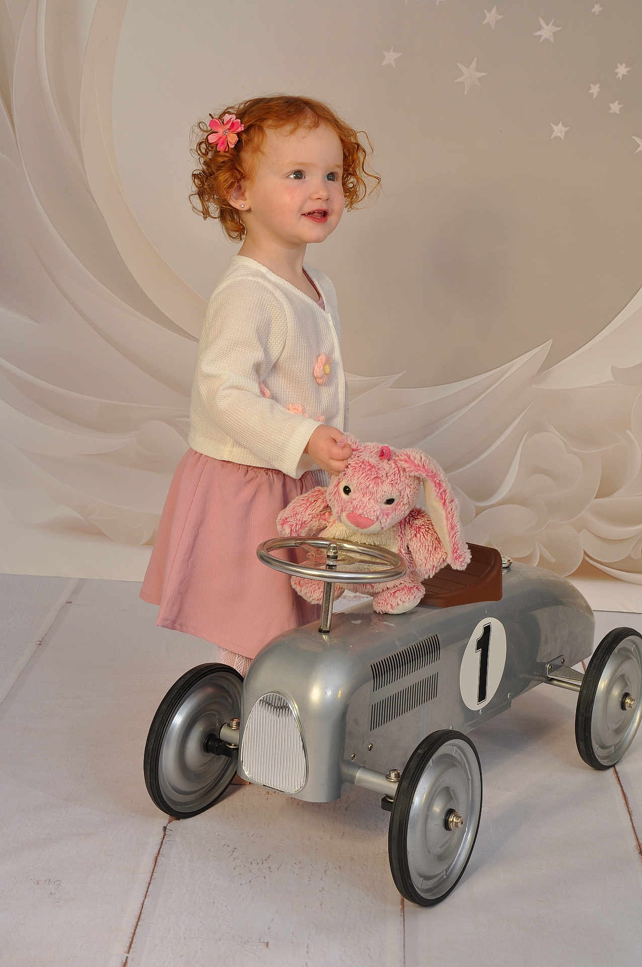 Aliya a rejoint le concours — aidez-le/la à gagner de superbes lots ! child, toddler, red_hair, curly_hair, flower_clip, pink_skirt, white_cardigan, toy_car, plush_bunny, indoor, floor, wheels, steering_wheel, cute, smiling, portrait, studio, decor, stars, play