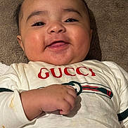 Zohan is registered to the contest to win money with this photo: apparel, baby, baby_girl, blanket, clothing, cute, eyelashes, face, gucci, hand, happy, indoors, infant, lying_down, person, portrait, skin, smiling, warm, young_child