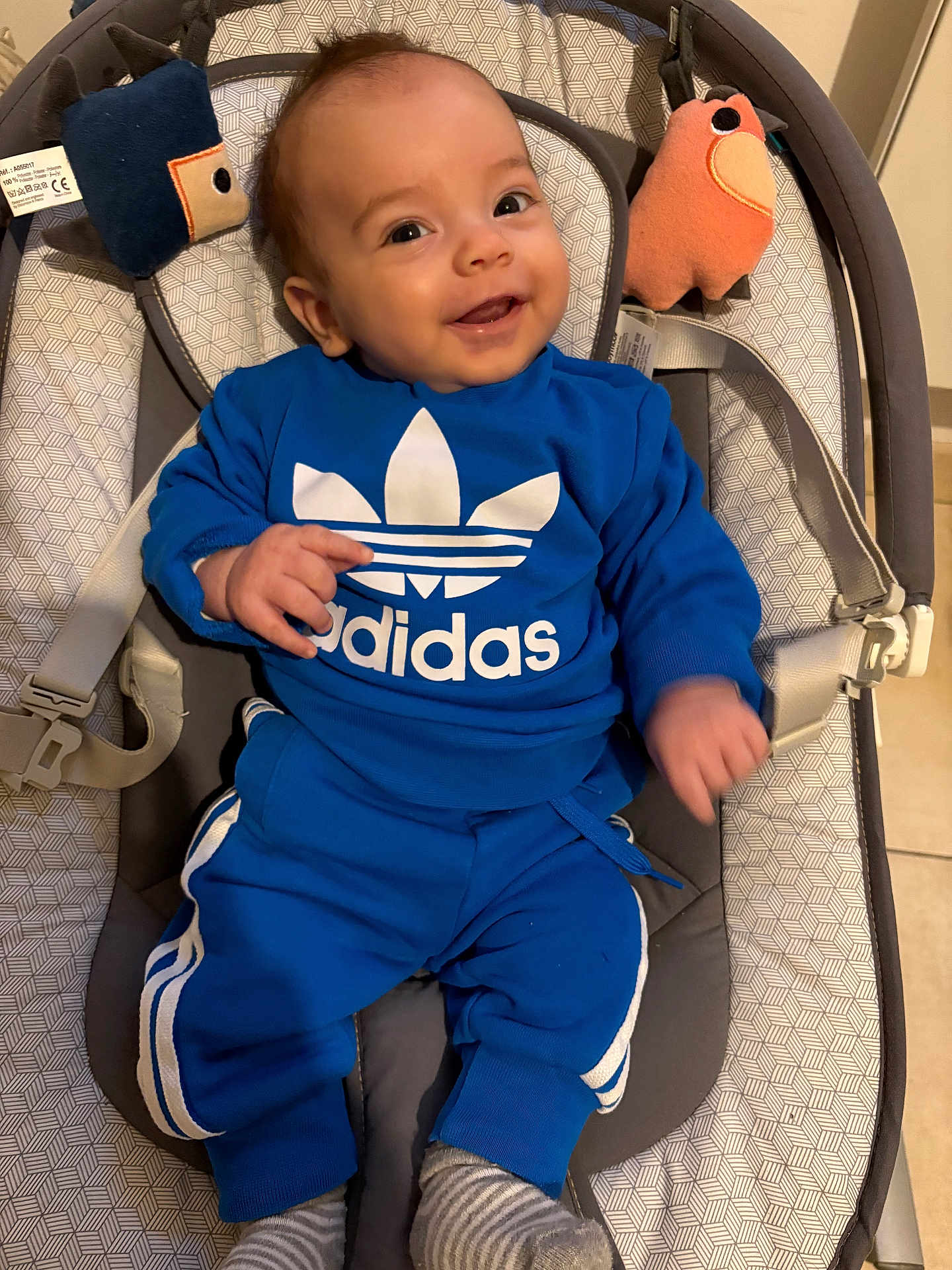 Noé participe au concours pour gagner de l'argent avec cette photo : baby, child, smile, blue_clothing, adidas, plush_toy, baby_seat, infant, happy, indoors, cushion, footwear, striped_socks, person, portrait, cute, comfortable, cozy, pointing, young_child