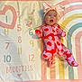baby, infant, onesie, headband, bow, milestone_blanket, numbers, colorful, pink, red, lying_down, cute, child, apparel, patterned_clothing, expression, indoors, soft_texture, pastel_colors, playtime