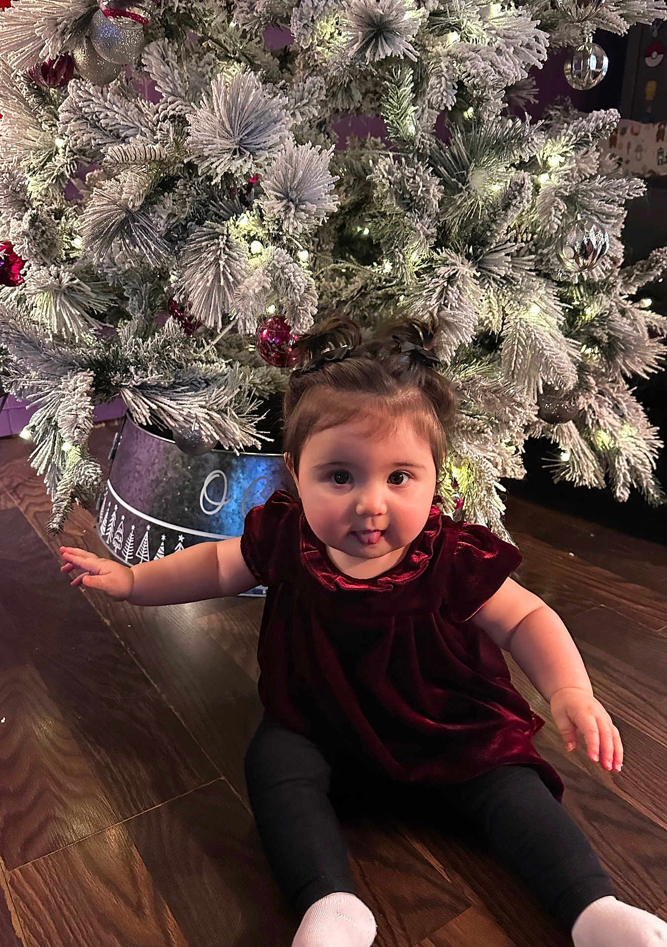 Novalee is registered to the contest to win money with this photo: baby, child, christmas_tree, holiday, indoor, floor, wood_floor, decorations, ornaments, lights, festive, cute, playful, pigtails, velvet_dress, black_leggings, tongue_out, sitting, holiday_season, smiling