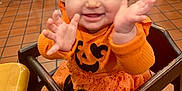 Novalee is registered to the contest to win money with this photo: baby, child, high_chair, orange_clothing, pumpkin_outfit, smiling, happy, clapping_hands, indoor, restaurant, wooden_furniture, toddler, cute, person, face, hair_tie, black_socks, tile_floor, table, chair