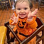 baby, child, high_chair, orange_clothing, pumpkin_outfit, smiling, happy, clapping_hands, indoor, restaurant, wooden_furniture, toddler, cute, person, face, hair_tie, black_socks, tile_floor, table, chair