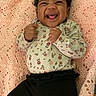 Ameera is registered to the contest to win money with this photo: baby, smiling, happy, infant, curly_hair, blanket, pink, crocheted, floral, patterned_clothing, long_sleeve, black_pants, lying_down, indoors, cute, joyful, person, child, portrait, fist_raised
