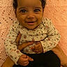 baby, smiling, infant, hands, knitted_blanket, pink, cute, child, portrait, happy, clothing, long_sleeve, black_pants, curly_hair, skin, person, indoor, holding, young, seated