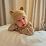 baby, infant, child, portrait, close_up, wide_eyed, surprised, smiling, bear_hat, knit_sweater, beige_clothing, bed, blanket, bedroom, curtains, indoors, soft_lighting, cozy, sitting, adorable